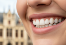 Close-up of a bright, healthy smile with perfectly contoured pink gums and sparkling white teeth, set against a softly blurred, neutral background suggesting a European city street in Belgium.