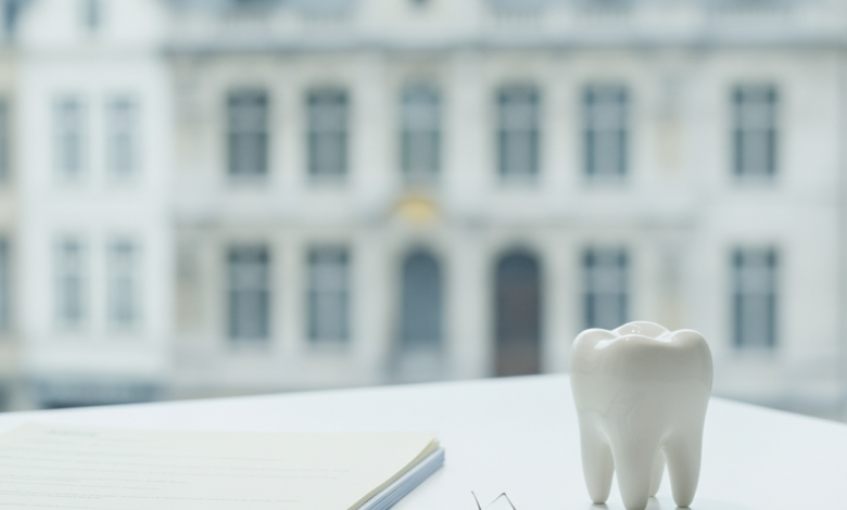 A photorealistic image showing a stack of generic insurance documents next to a clean, minimalist dental model on a modern table. In the blurred background, a classic Belgian architectural building suggests the location. The scene represents healthcare finance and dental reimbursement.