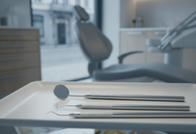 A photorealistic close-up of sterile dental instruments on a clean white tray in the foreground. The background is softly blurred, showing abstract shapes that suggest scattered euro coins and paper bills, conceptually representing hidden financial costs. Behind these, a very indistinct urban European street scene is visible through a window, implying a location like Belgium. The lighting is clean and professional.